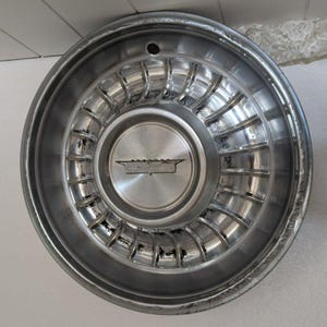 Puede incluir: Un tapacubos de coche plateado con un logotipo de Cadillac en el centro. El tapacubos tiene un diseño radial con una serie de radios que irradian desde el centro.