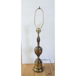 Puede incluir: Una lámpara de pie de latón vintage con un diseño decorativo de piña en el tallo. La lámpara tiene una base redonda y un brazo curvo que sostiene una bombilla. La lámpara es de color latón cálido y está enchufada a una toma de corriente.