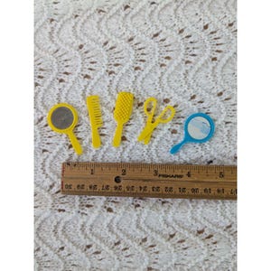 May include: A collection of miniature yellow and blue grooming tools, including a comb, brush, scissors, and mirrors. The ruler next to the tools measures approximately 5 inches in length. The tools are arranged on a white, textured surface.
