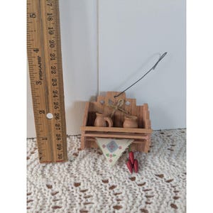 May include: A miniature wooden shelf with two small jugs, a patterned cloth, and red chili peppers. The shelf is approximately 3 inches wide and has a string for hanging. A ruler is next to the shelf.