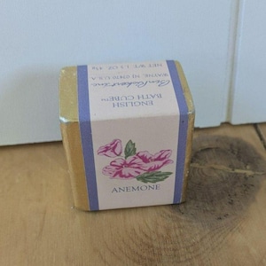 May include: A gold-coloured English bath cube with a pink and purple label. The label features a pink anemone flower illustration and the word "ANEMONE". The cube weighs 45g.