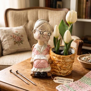 May include: A ceramic figurine of an elderly person in a rocking chair next to a woven-textured planter containing three white tulips. The figurine wears glasses and a pink outfit. A tag reads Grandma's Knitting Corner. Two knitting needles lie nearby.