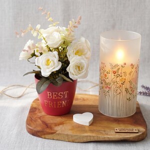 Peut inclure: Un pot rouge avec le texte BEST FRIEND en doré, contenant des roses artificielles blanches, à côté d'un photophore cylindrique en verre dépoli avec un motif floral et une lumière chaude, posés sur une planche en bois avec un petit cœur blanc.