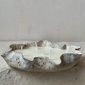 Könnte beinhalten: Eine große dekorative Kerze in einer gesprenkelten, cremefarbenen Keramikschale. Die Schale hat eine einzigartige, organische Form mit gewelltem Rand. Die Kerze hat cremefarbenes Wachs und mehrere Dochte.