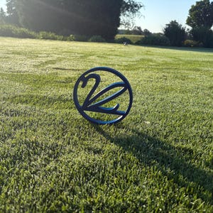 May include: A blue metal golf course marker with the number 2 inside a circle, set on a green grassy field. The marker casts a shadow in the sunlight. The background features more green grass and trees.