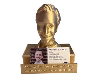 May include: A gold-colored bust of a person with a business card holder. The business card holder has a business card with the name Andrew Buchko, Realtor, and contact information. The business card holder also has a smaller plaque with the name Andrew Buchko and the phone number (734) 735-4182.