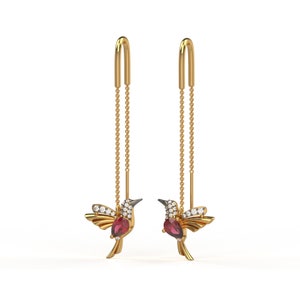 Puede incluir: Un par de pendientes colgantes de oro con un diseño de colibrí. Cada colibrí tiene una piedra preciosa roja y piedras preciosas transparentes. Los pendientes tienen una cadena que cuelga de la parte superior de la oreja.