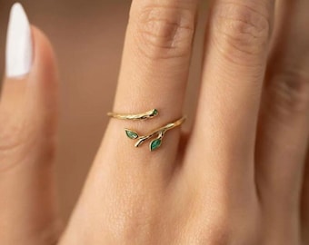 May include: A gold ring with a delicate branch design and green leaves. The ring is open and wraps around the finger.