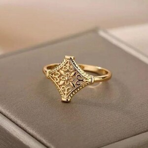 Op de afbeelding: Een gouden ring met een vierkant ontwerp met ingewikkelde bloem- en geometrische patronen.