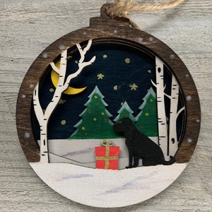 May include: A wooden Christmas ornament with a layered design. The ornament depicts a snowy forest scene with a crescent moon, two pine trees, a dog, and a red gift box. The ornament is painted in shades of brown, white, green, blue, and yellow.