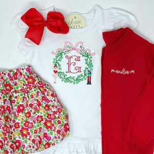 Puede incluir: Un conjunto infantil con una camiseta blanca con bordado de corona e inicial, una falda floral y una rebeca roja. También se incluye un gran lazo rojo y una etiqueta que dice "three bees". El conjunto está diseñado para un niño.