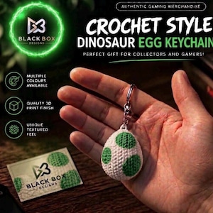 May include: A white egg-shaped keyring with green spots, featuring a textured surface that mimics a crochet pattern. The keyring is attached to a metal ring. Text on the image includes "CROCHET STYLE DINOSAUR EGG KEYCHAIN" and "BLACK BOX DESIGNS".