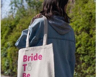 Op de afbeelding: Een witte canvas tote bag met roze tekst die "Bride To Be" zegt en een diamanten ring illustratie.