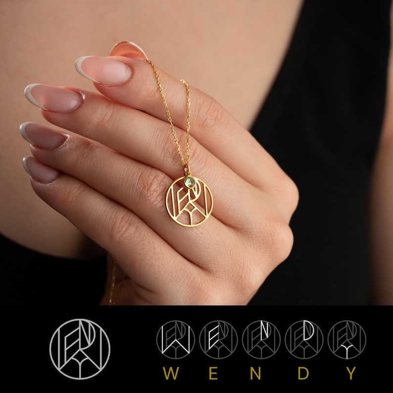 May include: Gold necklace with a circular pendant featuring a geometric design and a small green gemstone. The pendant is inscribed with the letters "W", "E", "N", "D", and "Y".
