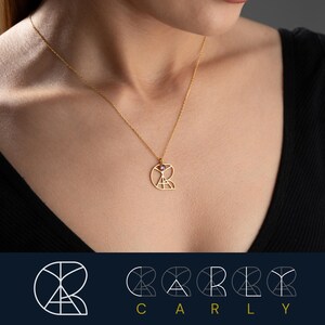 May include: A gold necklace with a geometric pendant featuring the letter 'R' with a small pink gemstone in the center. The pendant is made of thin gold wire and has a delicate, intricate design. The necklace is displayed on a model wearing a black top.