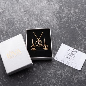 May include: A set of gold jewelry featuring a necklace and earrings. The necklace and earrings have a geometric design with a pink stone in the center. The jewelry is packaged in a white box with the brand name "ICVA Jewelry" printed on it. The box is sitting on a gray surface. A white card with the brand name "CARLY" printed on it is also on the surface.