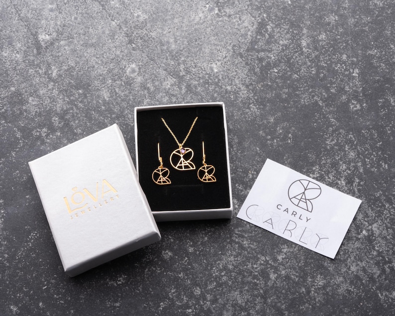 May include: A set of gold jewelry featuring a necklace and earrings. The necklace and earrings have a geometric design with a pink stone in the center. The jewelry is packaged in a white box with the brand name "ICVA Jewelry" printed on it. The box is sitting on a gray surface. A white card with the brand name "CARLY" printed on it is also on the surface.