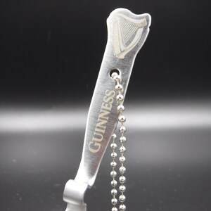 May include: A silver-colored bottle opener with the Guinness logo and text. The opener has a harp design at the top and a ball chain attached. The background is dark.