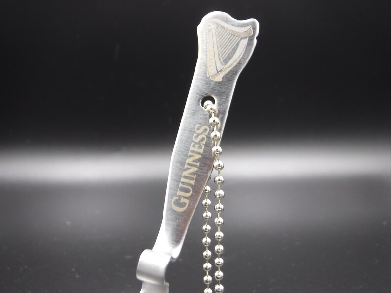 May include: A silver-colored bottle opener with the Guinness logo and text. The opener has a harp design at the top and a ball chain attached. The background is dark.