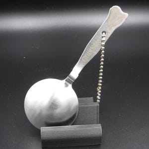 May include: A stainless steel spoon with a round bowl and a handle shaped like a bone. The handle is attached to a small ball chain. The spoon is resting on a black stand. The word "GUINNESS" is etched on the handle.