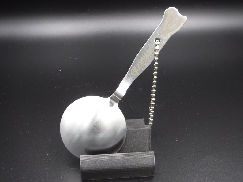 May include: A stainless steel spoon with a round bowl and a handle shaped like a bone. The handle is attached to a small ball chain. The spoon is resting on a black stand. The word "GUINNESS" is etched on the handle.