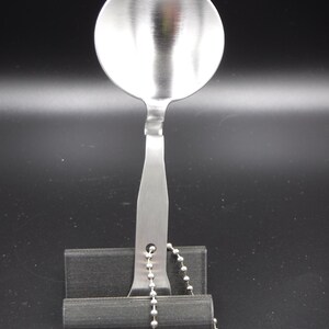 May include: A stainless steel spoon with a round bowl and a long handle. The handle has a hole and is attached to a silver ball chain. The spoon is resting on a black stand against a black background.
