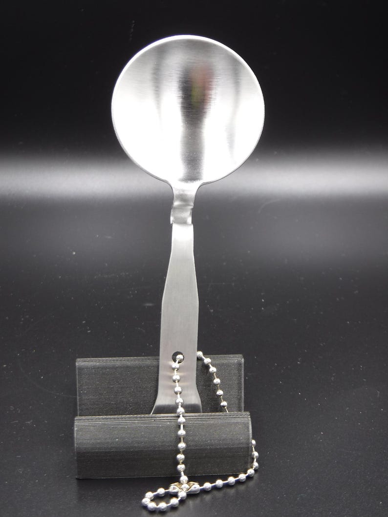 May include: A stainless steel spoon with a round bowl and a long handle. The handle has a hole and is attached to a silver ball chain. The spoon is resting on a black stand against a black background.