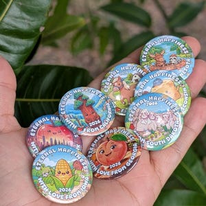 Puede incluir: Una colección de chapas redondas con coloridas ilustraciones de dibujos animados de personajes y monumentos estatales, cada una etiquetada con Eternal Happiness, el año 2026 y el nombre del estado como Iowa, Georgia, California y Dakota del Sur.