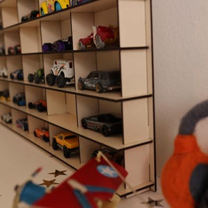 May include: A wooden shelf unit with multiple compartments, designed to hold and display toy cars. The shelf is made of light brown wood and has a natural finish. The shelf is filled with various toy cars, including red, blue, yellow, and black cars.