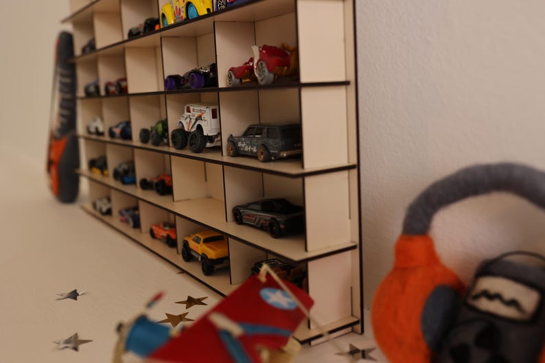 May include: A wooden shelf unit with multiple compartments, designed to hold and display toy cars. The shelf is made of light brown wood and has a natural finish. The shelf is filled with various toy cars, including red, blue, yellow, and black cars.