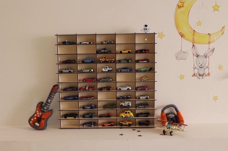 May include: A wooden toy car display shelf with multiple compartments, filled with toy cars. The shelf is mounted on a white wall. There is a toy guitar and a toy airplane on the floor in front of the shelf.