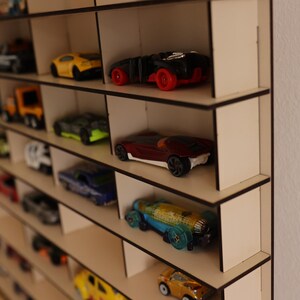 May include: A wooden shelf unit with multiple shelves, each holding a collection of toy cars. The cars are in various colors, including red, blue, yellow, and black. The shelf unit is made of light wood and has a simple design.