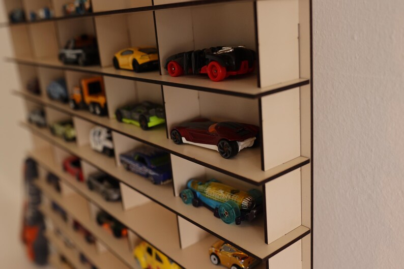May include: A wooden shelf unit with multiple shelves, each holding a collection of toy cars. The cars are in various colors, including red, blue, yellow, and black. The shelf unit is made of light wood and has a simple design.