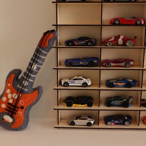 May include: A wooden shelf unit with multiple shelves, each holding a different toy car. The cars are in various colors, including red, blue, green, and black. A felt orange and gray electric guitar is leaning against the shelf unit.