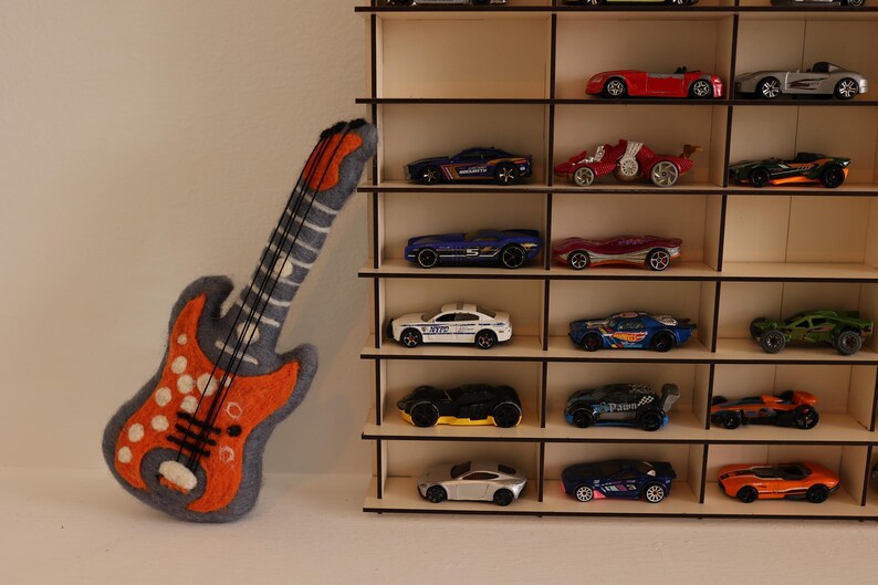 May include: A wooden shelf unit with multiple shelves, each holding a different toy car. The cars are in various colors, including red, blue, green, and black. A felt orange and gray electric guitar is leaning against the shelf unit.