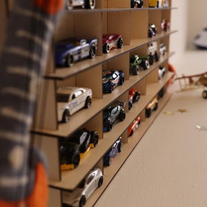 May include: A wooden toy car display shelf with multiple levels. The shelf is filled with toy cars in various colors, including red, blue, black, and silver.
