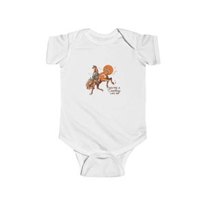 May include: White baby bodysuit featuring a cartoon illustration of a cowboy riding a bucking horse. The text "You're a Cowboy Like Me" is printed below the image.