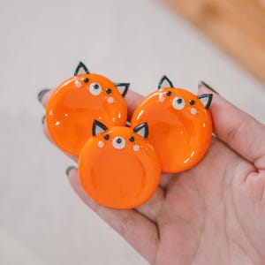 May include: Three orange, round, fox-shaped decorative items with black ears and white accents. The items are glossy and appear to be made of resin or a similar material. The fox faces have small black eyes and white dots.
