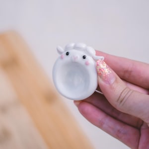 Può includere: Un piccolo oggetto in ceramica bianco e circolare a forma di testa di pecora con un centro cavo. La pecora presenta occhi neri, un naso rosa e piccole guance rosa, con una texture di lana bianca scolpita sulla parte superiore della testa.