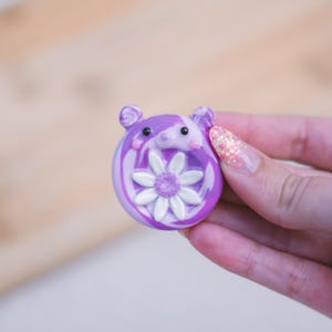 Può includere: Un ciondolo circolare a forma di orso, marmorizzato nei colori viola e bianco, con un fiore di margherita bianco dal centro testurizzato sulla parte anteriore. L'orso presenta due piccole orecchie arrotondate, due occhi neri e un naso rosa.