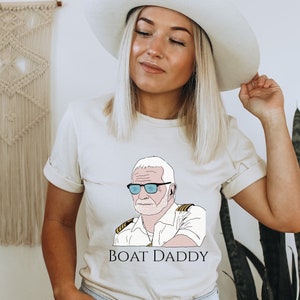 May include: A beige t-shirt with a cartoon illustration of a man wearing sunglasses and a captain's hat. The text "BOAT DADDY" is printed below the illustration.