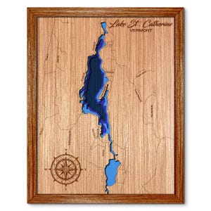 May include: A framed wooden bathymetric map of Lake St. Catherine, Vermont. The map features layered blue water depths set into a light wood grain surface with engraved road names, a compass rose in the bottom left, and the text Lake St. Catherine.