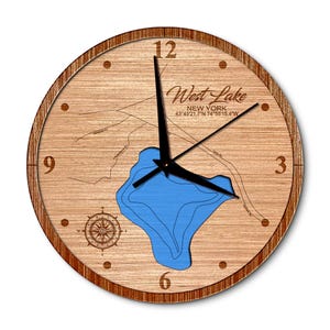 May include: A round wooden wall clock featuring a map of West Lake, New York, with a blue lake outline. The clock face has black hands, a compass rose, and the text "West Lake NEW YORK 43°45'21.7"N 74°55'15.4"W". The clock has a dark brown wooden frame.