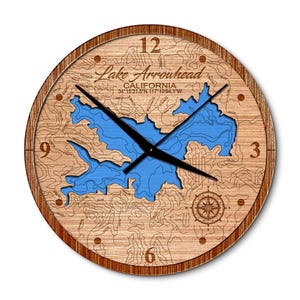 May include: A wooden clock with a map of Lake Arrowhead, California. The clock face is surrounded by a brown wooden frame. The map shows the lake's outline and surrounding terrain. The clock face features Roman numeral hour markers and black hands.