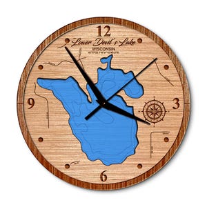 May include: A round wooden clock featuring a blue lake map of Lower Devil's Lake, Wisconsin. The clock has black hands, a compass rose, and the numbers 3, 6, 9, and 12. The clock's outer rim is a darker brown.