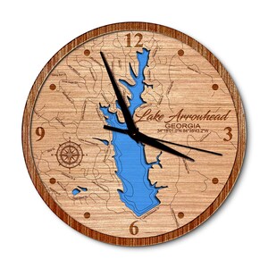 May include: A round wooden wall clock featuring a laser-engraved map of Lake Arrowhead, Georgia. The center displays a bright blue cutout of the lake, surrounded by detailed street names, a compass rose, and coordinates 34°18'01.0"N 84°35'43.2"W.