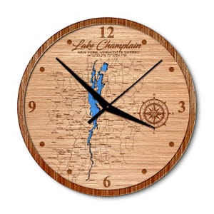 May include: A round wooden wall clock featuring a laser-engraved map of Lake Champlain, New York, Vermont, and Quebec. The clock face displays the lake in blue, a compass rose, coordinates, and black hour and minute hands with numerals 3, 6, 9, 12.