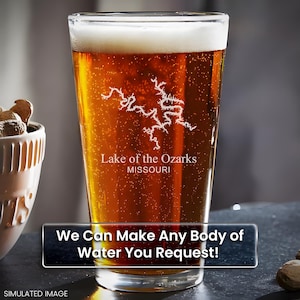 May include: A clear glass pint glass with a frosted design of Lake of the Ozarks, Missouri. The glass is filled with a golden brown beer. The text "We Can Make Any Body of Water You Request!" is visible at the bottom of the image.