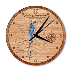 May include: A wooden clock with a map of Lake Champlain, New York, Vermont, and Quebec. The clock face features the lake's outline in blue, with the text "Lake Champlain" and coordinates "44°32′33.2″N 73°19′54.1"W".