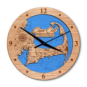 May include: A round wooden clock featuring a detailed map of Cape Cod, Massachusetts, with a blue ocean background. The clock face has black hands, a compass rose, and the text "Cape Cod, Massachusetts". The clock is approximately 12 inches in diameter.